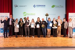 Gruppenfoto auf einer Bühne: Mehrere Teilnehmende stehen nebeneinander, halten Urkunden und Blumensträuße in den Händen und heben teilweise die Arme. Im Hintergrund ist eine Projektionsfläche mit der Überschrift „Herzlichen Glückwunsch!“ und Logos beteiligter Unternehmen zu sehen.