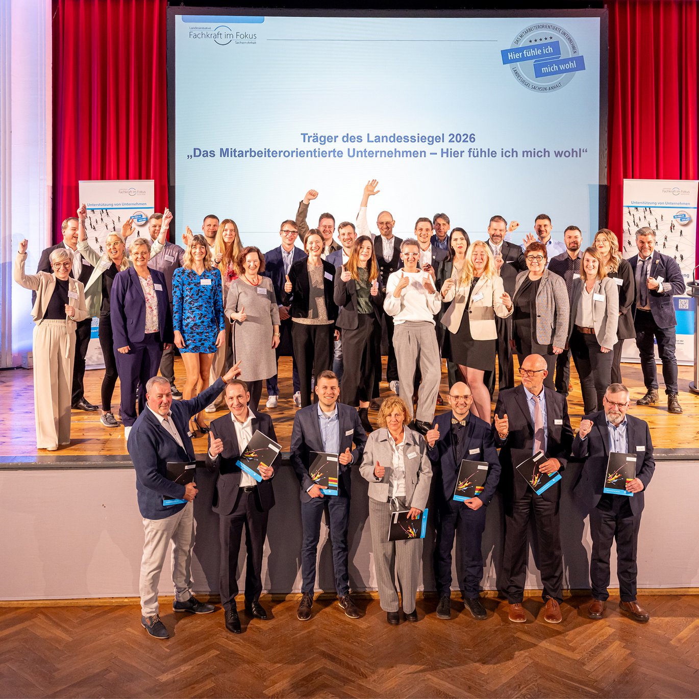 Gruppenfoto der Träger des Landessiegels 2026 „Das mitarbeiterorientierte Unternehmen – Hier fühle ich mich wohl“ aus Sachsen‑Anhalt bei der feierlichen Auszeichnung auf einer Bühne.