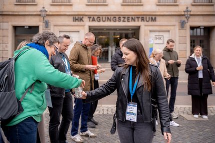 Eine Gruppe Menschen steht vor dem Eingang eines Gebäudes mit der Aufschrift „IHK Tagungszentrum“. Mehrere Personen tragen Namensschilder und halten kleine Zettel in der Hand.
