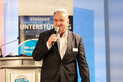 Redebeitrag bei einer Unternehmensveranstaltung: Eine Person steht neben einem Rednerpult und spricht in ein Mikrofon; im Hintergrund ist ein Roll‑up‑Banner mit dem Schwerpunkt Strategie und Unterstützung zu sehen.