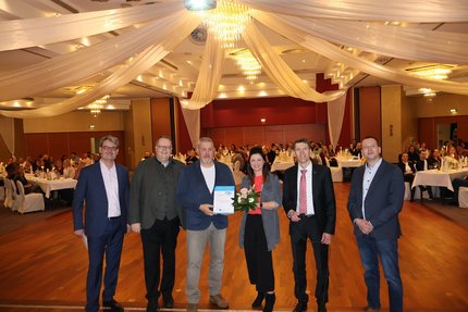 Eine Gruppe Menschen stehen in einem Festsaal auf der Tanzfläche. Im Hintergrund sitzen Menschen an festlichen Tafeln.