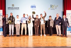 Gruppenfoto auf einer Bühne: Mehrere Teilnehmende stehen nebeneinander, halten Urkunden und Blumensträuße in den Händen und heben teilweise die Arme. Im Hintergrund ist eine Projektionsfläche mit der Überschrift „Herzlichen Glückwunsch!“ und Logos beteiligter Unternehmen zu sehen.