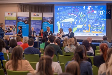 Viele Menschen sitzen in Stuhlreihen und schauen auf ein Podium, bestehend aus fünf Personen.