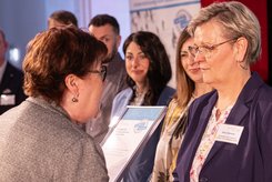 Übergabe einer Urkunde bei einer Veranstaltung: Zwei Personen stehen im Vordergrund und schütteln sich die Hände, während eine Urkunde überreicht wird; im Hintergrund stehen weitere Teilnehmende in einer Reihe auf der Bühne.