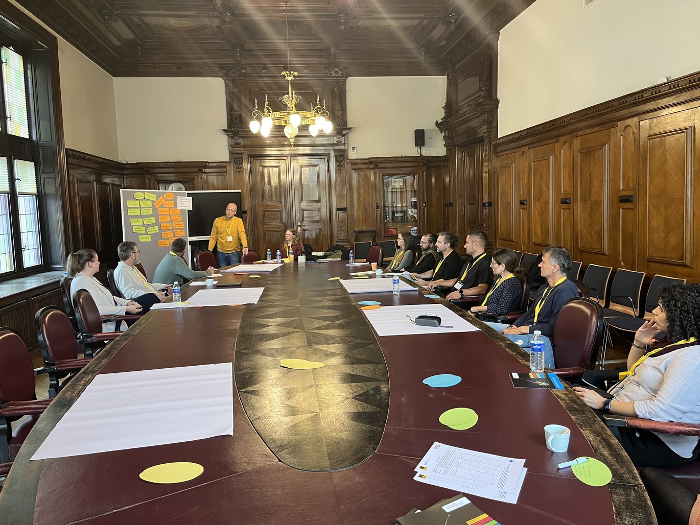 12 Personen sitzen bzw. stehen um einen großen Tisch in einem holzvertäfelten Raum, auf dem Tisch liegen Workshopmaterialien, daneben steht eine mit bunten Zetteln bestückte Moderationswand