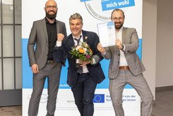 Drei Teilnehmende stehen vor einem Banner zum Landessiegel „Hier fühle ich mich wohl“ in Sachsen‑Anhalt. Eine Person hält eine Urkunde in die Kamera, eine weitere präsentiert einen Blumenstrauß; alle tragen formelle Kleidung und posieren für ein Foto.