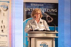 Redebeitrag bei einer Veranstaltung: Eine Person steht hinter einem Rednerpult und spricht in ein Mikrofon; hinter dem Podium ist ein Banner mit Begriffen wie Strategie und Unterstützung zu sehen.