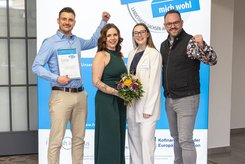 Vier Teilnehmende stehen vor einem Banner zum Landessiegel „Hier fühle ich mich wohl“ in Sachsen‑Anhalt. Eine Person hält eine Urkunde in die Kamera, eine weitere präsentiert einen Blumenstrauß; alle tragen formelle Kleidung und posieren für ein Foto.