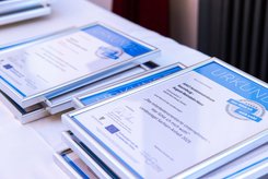 Mehrere gerahmte Urkunden liegen gestapelt auf einem Tisch; die Auszeichnungen tragen das Landessiegel „Hier fühle ich mich wohl“ und sind für mitarbeiterorientierte Unternehmen bestimmt.