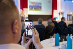 Blick aus dem Publikum auf eine Veranstaltung: Eine Person hält ein Smartphone und fotografiert eine Rede auf der Bühne; auf dem Display ist das Rednerpult zu sehen. 
