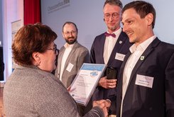 Zwei Personen schütteln sich die Hände, während eine Urlkunde übergeben wird. Daneben stehen weitere Personen und schauen zu.