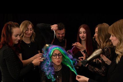 Eine Gruppe Menschen stehen um einen Menschen mit grün-lila Haaren und frisieren diese.