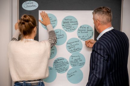 Zwei Menschen stehen an einer Pinnwand und befestigen ein gestaltetes Chart.