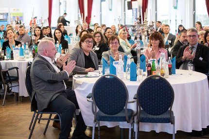 Großer Veranstaltungssaal mit vielen Teilnehmenden an runden, gedeckten Tischen; die Gäste sitzen und stehen in Gesprächsrunden.