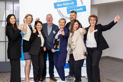 Acht Personen stehen nebeneinander vor einem blau-weißen Messestand mit dem Schriftzug des Landessiegel „Hier fühle ich mich wohl“. 