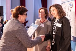 Urkundenübergabe auf einer Bühne: Eine Person überreicht einer anderen Person eine gerahmte Urkunde; daneben steht eine weitere Teilnehmende Person und schaut zu.