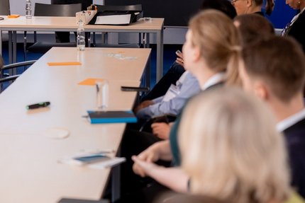 eine Gruppe Menschen sitzen am Tisch und schauen nach vorn zum Referenten.