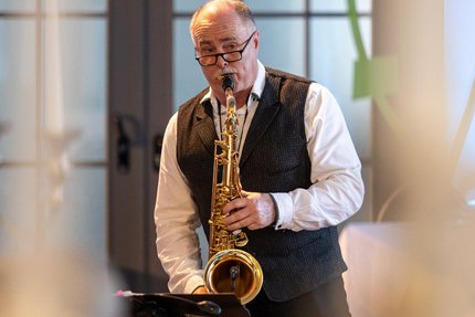 Musizierende Person spielt ein Saxophon in einem Veranstaltungsraum; im Vordergrund ist das Instrument zu sehen, im Hintergrund befinden sich Kerzenleuchter und unscharf dargestellte Gäste.