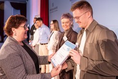 Eine Person überreicht einer anderen eine Urkunde; daneben stehen weitere Teilnehmende und schauen zu