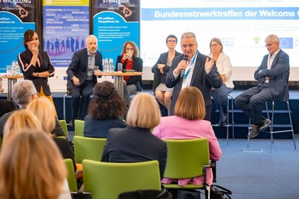 Fünf Menschen sitzen auf Barhockern vor einer Bühne. Zwei Menschen stehen davor und moderieren.