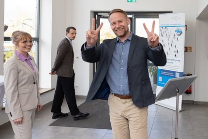 Teilnehmende stehen neben einem Roll-up-Banner; eine Person im Vordergrund zeigt ein V‑Zeichen.