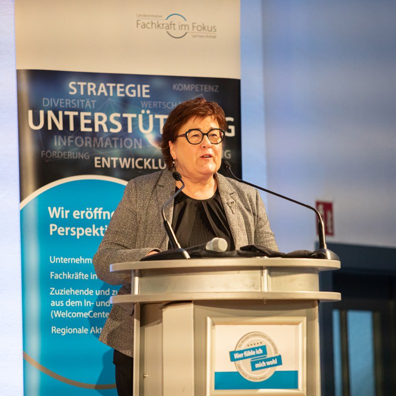 Redebeitrag bei einer Veranstaltung: Eine Person steht hinter einem Rednerpult und spricht in ein Mikrofon; hinter der Person ist ein Banner mit Begriffen wie Strategie, Unterstützung und Entwicklung zu sehen.