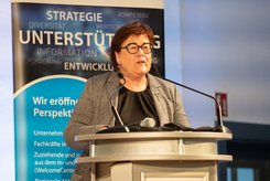 Redebeitrag bei einer Veranstaltung: Eine Person steht hinter einem Rednerpult und spricht in ein Mikrofon; hinter der Person ist ein Banner mit Begriffen wie Strategie, Unterstützung und Entwicklung zu sehen.