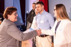 Übergabe einer Urkunde bei einer Veranstaltung: Zwei Personen stehen im Vordergrund und schütteln sich die Hände, während eine Urkunde überreicht wird; im Hintergrund stehen weitere Teilnehmende in einer Reihe auf der Bühne.