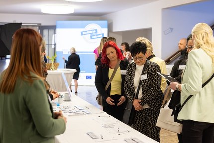 Mehrere Teilnehmende stehen an einem Registrierungstisch in einem hellen Veranstaltungsfoyer und sehen Namensschilder und Unterlagen durch; im Hintergrund ist ein Veranstaltungsbanner zu sehen.
