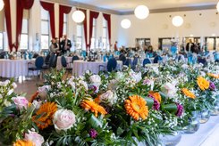 Viele Blumensträuße nebeneinander auf einerm Tisch. Im Hintergrund ist ein großer Saal festlich geschmückt.