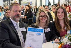 Teilnehmende am Tisch präsentieren eine Urkunde bei einer Festveranstaltung; im Hintergrund sitzen weitere Gäste im Saal.