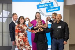 Sechs Personen stehen nebeneinander vor einem blau-weißen Messestand mit dem Schriftzug des Landessiegel „Hier fühle ich mich wohl“. 
