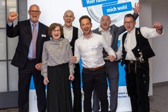 Sechs Personen stehen nebeneinander vor einem blau-weißen Messestand mit dem Schriftzug des Landessiegel „Hier fühle ich mich wohl“. 