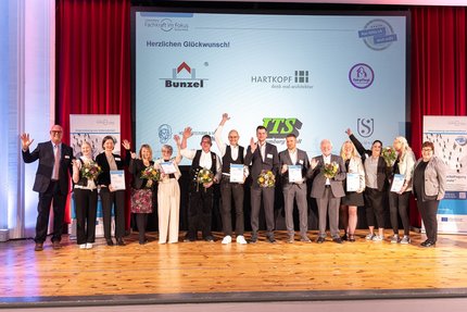 Gruppenfoto auf einer Bühne: Mehrere Teilnehmende stehen nebeneinander, halten Urkunden und Blumensträuße in den Händen und jubeln. Im Hintergrund ist eine Projektionsfläche mit der Überschrift „Herzlichen Glückwunsch!“ und Logos beteiligter Unternehmen zu sehen.