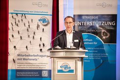 Redebeitrag bei einer Veranstaltung: Eine Person steht hinter einem Rednerpult und spricht in ein Mikrofon; hinter dem Podium ist ein Banner mit Begriffen wie Strategie, Unterstützung und Entwicklung zu sehen.