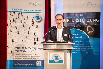 Redebeitrag bei einer Veranstaltung: Eine Person steht hinter einem Rednerpult und spricht in ein Mikrofon; hinter dem Podium ist ein Banner mit Begriffen wie Strategie, Unterstützung und Entwicklung zu sehen.