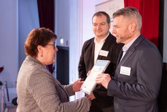 Urkundenübergabe auf einer Bühne: Eine Person überreicht einer anderen Person eine gerahmte Urkunde; daneben steht ein weiterer Teilnehmender und schaut zu.