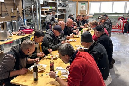 eine Gruppe Menschen sitzen in einer Werkhalle an einer langen Tafel und essen