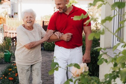 Ein jüngerer Mann begleitet eine ältere Frau durch ihren Garten.