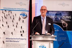 Redebeitrag bei einer Veranstaltung: Eine Person steht hinter einem Rednerpult und spricht in ein Mikrofon; hinter dem Podium ist ein Banner mit Begriffen wie Strategie, Unterstützung und Entwicklung zu sehen.