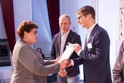 Eine Person übergibt einer anderen eine Urkunde; daneben stehen weitere Teilnehmende und schauen zu