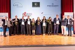 Gruppenfoto auf einer Bühne: Mehrere Teilnehmende stehen nebeneinander, halten Urkunden und Blumensträuße in den Händen und heben teilweise die Arme. Im Hintergrund ist eine Projektionsfläche mit der Überschrift „Herzlichen Glückwunsch!“ und Logos beteiligter Unternehmen zu sehen.