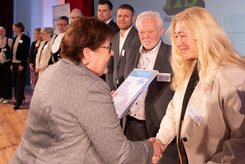 Übergabe einer Urkunde bei einer Veranstaltung: Zwei Personen stehen im Vordergrund und schütteln sich die Hände, während eine Urkunde überreicht wird; im Hintergrund stehen weitere Teilnehmende in einer Reihe auf der Bühne.