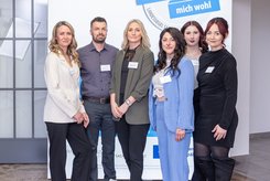 Sechs Personen stehen nebeneinander vor einem blau-weißen Messestand mit dem Schriftzug des Landessiegel „Hier fühle ich mich wohl“. 