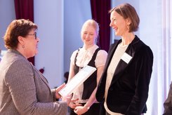 Urkundenübergabe auf einer Bühne: Eine Person überreicht einer anderen Person eine gerahmte Urkunde; daneben steht eine weitere teilnehmende Person und schaut zu.