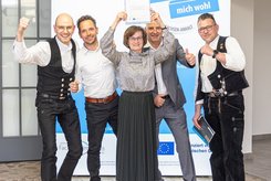 Fünf Teilnehmende posieren vor einem Banner zum Landessiegel „Das mitarbeiterorientierte Unternehmen – Hier fühle ich mich wohl“ in Sachsen‑Anhalt. Eine Person hält eine Urkunde über dem Kopf, während die anderen mit erhobenen Armen und Daumen nach oben für ein Gruppenfoto stehen.