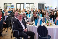 Großer Veranstaltungssaal mit zahlreichen Teilnehmenden an runden, gedeckten Tischen. Die Gäste sitzen und blicken nach vorne; auf den Tischen stehen Getränke, Unterlagen, blaue Servietten und Kerzenleuchter.
