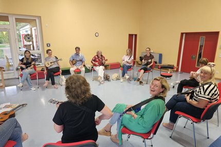 Eine Gruppe Menschen sitzen im Stuhlkreis und alle halten eine Gitarre in der Hand