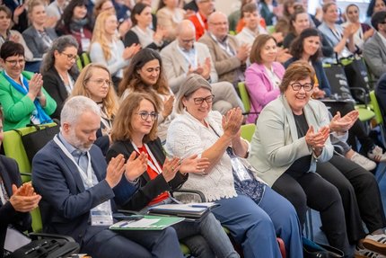 Viele Menschen sitzen in Stuhlreihen in einem großen Raum und klatschen in die Hände.