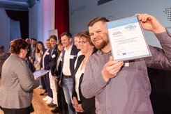 Person im Vordergrund hält eine gerahmte Urkunde in die Kamera; im Hintergrund stehen weitere Teilnehmende in einer Reihe auf einer Bühne, während eine Urkundenübergabe stattfindet.